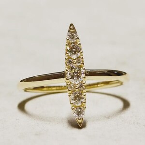 May include: A gold ring featuring a vertical marquise-shaped setting adorned with five round clear stones. The band is thin and polished, intersecting the centre of the vertical stone arrangement, creating a balanced and symmetrical jewellery piece.