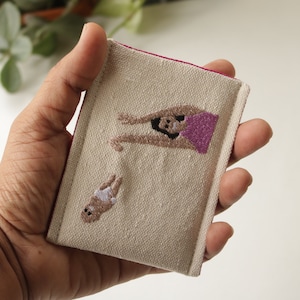 Puede incluir: Una peque&ntilde;a bolsa de tela beige con ribete rosa, que presenta im&aacute;genes bordadas de una mujer y un beb&eacute;. La mujer lleva una blusa rosa y tiene los brazos levantados en el aire. El beb&eacute; lleva un pa&ntilde;al blanco y tiene los brazos levantados en el aire.