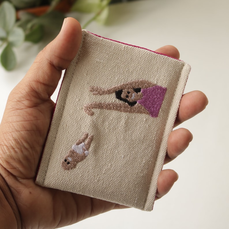 Puede incluir: Una peque&ntilde;a bolsa de tela beige con ribete rosa, que presenta im&aacute;genes bordadas de una mujer y un beb&eacute;. La mujer lleva una blusa rosa y tiene los brazos levantados en el aire. El beb&eacute; lleva un pa&ntilde;al blanco y tiene los brazos levantados en el aire.