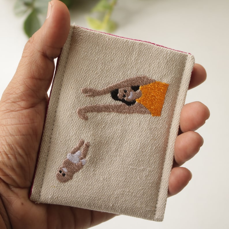 Puede incluir: Una cartera de lona beige con un borde rosa. La cartera tiene dos figuras bordadas. Una figura es una persona con una camiseta amarilla con los brazos levantados por encima de la cabeza. La otra figura es una persona con un mono blanco con los brazos y las piernas extendidos.
