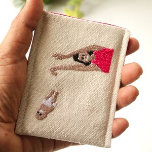 Puede incluir: Una bolsa de lona beige con ribete rosa presenta im&aacute;genes bordadas de una mujer en traje de ba&ntilde;o rojo y un beb&eacute; con pa&ntilde;al.