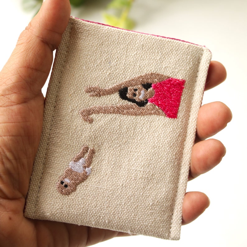 Puede incluir: Una bolsa de lona beige con ribete rosa presenta im&aacute;genes bordadas de una mujer en traje de ba&ntilde;o rojo y un beb&eacute; con pa&ntilde;al.