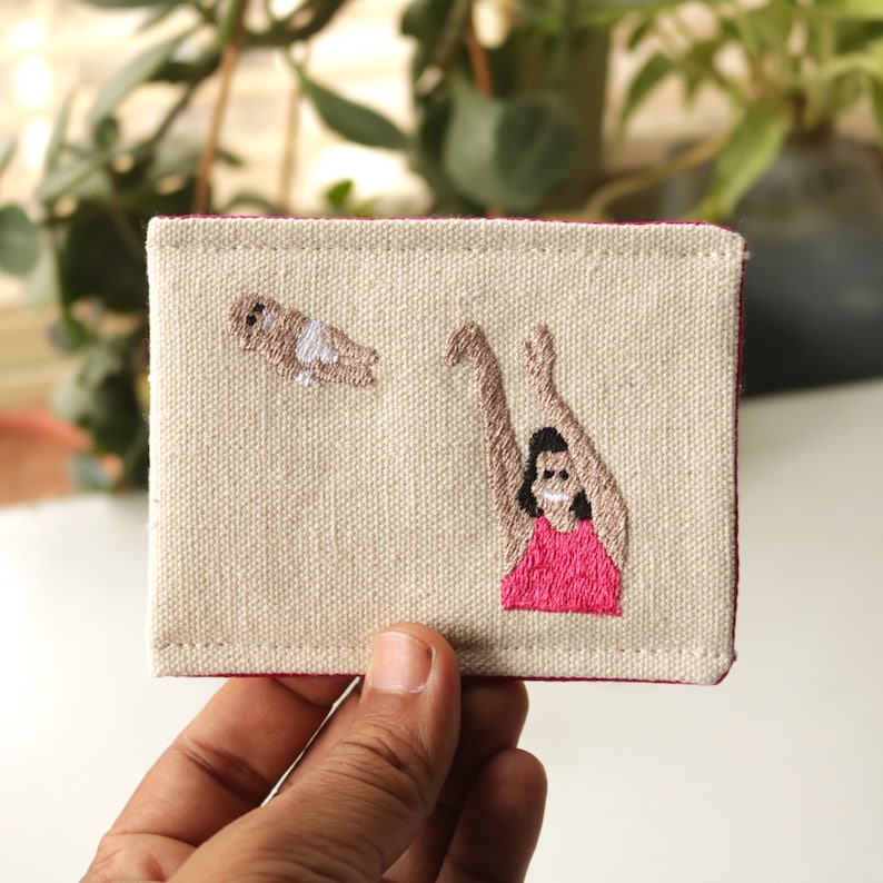 Puede incluir: Una bolsa de lona beige con costuras rosas presenta un dise&ntilde;o bordado de una persona con los brazos extendidos atrapando una peque&ntilde;a figura. La persona lleva una camisa rosa y la peque&ntilde;a figura lleva una camisa blanca.