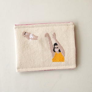 Puede incluir: Un porta tarjetas de lona blanca con costuras rosas. El porta tarjetas tiene una imagen bordada de una persona en un traje de ba&ntilde;o amarillo atrapando un beb&eacute; en el aire.