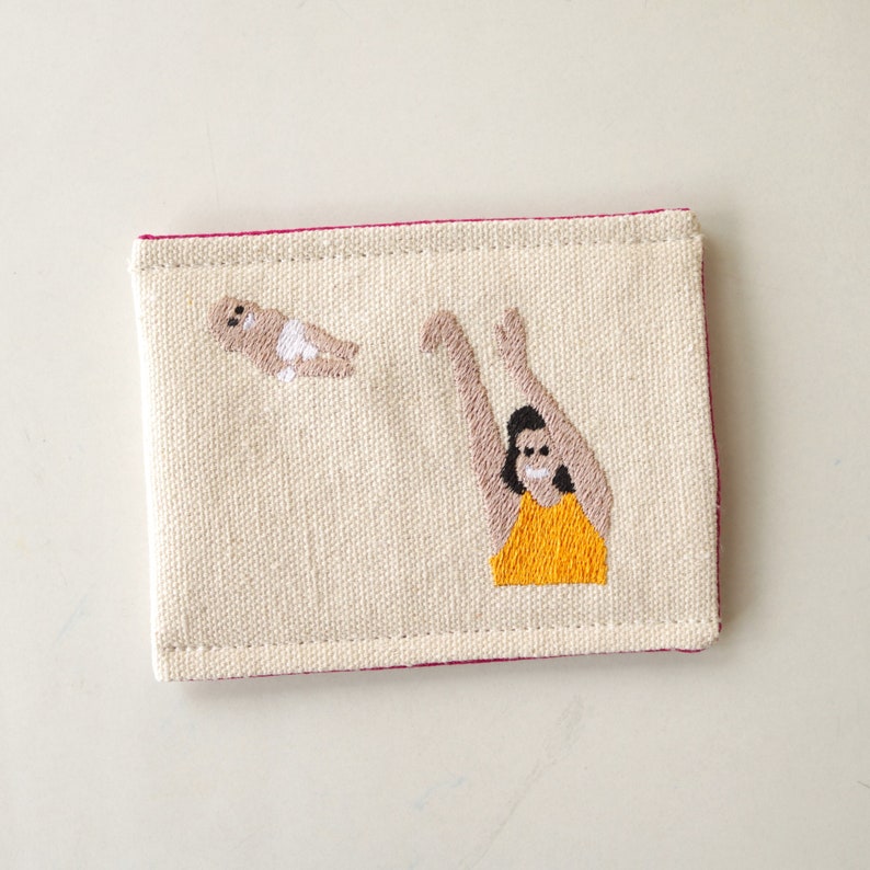 Puede incluir: Un porta tarjetas de lona blanca con costuras rosas. El porta tarjetas tiene una imagen bordada de una persona en un traje de ba&ntilde;o amarillo atrapando un beb&eacute; en el aire.