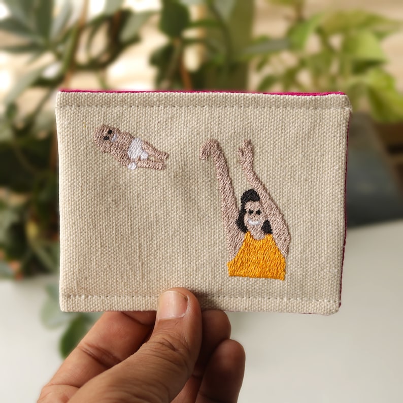 Puede incluir: Un porta tarjetas de lona beige con costuras rosas. El porta tarjetas presenta un dise&ntilde;o bordado de una persona con los brazos extendidos atrapando a una persona peque&ntilde;a que cae del cielo. La persona que atrapa a la persona que cae lleva una camisa amarilla.