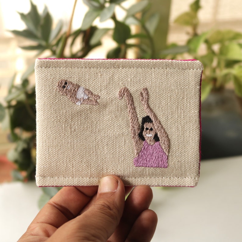 Puede incluir: Una bolsa de lona beige con costuras rosas presenta un dise&ntilde;o bordado de una persona con los brazos extendidos y una persona peque&ntilde;a volando sobre ella.
