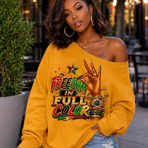 Puede incluir: Una sudadera de manga larga color amarillo mostaza con un estampado gráfico de una mano haciendo un gesto de "ok", adornada con pulseras doradas y un reloj. El texto dice "FREEDOM IN FULL COLOR" en rojo, verde y amarillo, con una estrella.