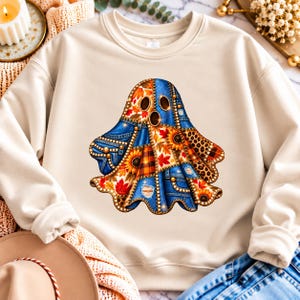 Könnte beinhalten: Ein beigefarbenes Sweatshirt mit Rundhalsausschnitt und einem zentralen Geistermotiv. Der Geist besteht aus einem Patchwork-Design mit blauem Jeansstoff, orangefarbenen und roten Herbstblättern, gelben Sonnenblumen, braunem Karomuster und Leopardenmuster.