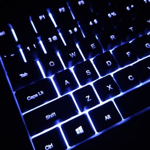 Może przedstawiać: Czarna klawiatura z białymi napisami i niebieskimi podświetlonymi klawiszami. Klawisze są oznaczone literami, cyframi i symbolami. Klawiatura jest podświetlona niebieskim światłem.