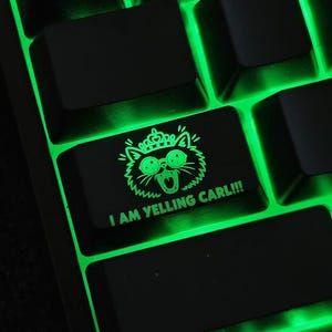 May include: A black mechanical keyboard keycap featuring a glowing green graphic of a cat wearing a crown and round glasses with its mouth open. The text I AM YELLING CARL!!! is printed in green below the cat illustration on the keycap surface.