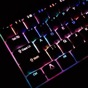 May include: Close-up of a black mechanical keyboard with illuminated keys. The keys are lit with a rainbow of colors, including red, blue, green, and purple. The keyboard has a compact layout with various symbols and letters.