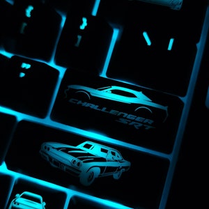 May include: A black keyboard with blue backlit keys. The keys have images of cars on them, including a Dodge Challenger SRT. The keyboard is lit up in the dark.