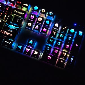 May include: A black keyboard with colorful backlit keys featuring Star Wars themed keycaps. The keys are labeled with numbers, letters, and symbols. The keycaps have various Star Wars characters and symbols, including the Millennium Falcon, Darth Vader, and the Death Star.