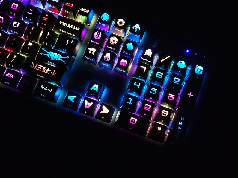 May include: A black keyboard with colorful backlit keys featuring Star Wars themed keycaps. The keys are labeled with numbers, letters, and symbols. The keycaps have various Star Wars characters and symbols, including the Millennium Falcon, Darth Vader, and the Death Star.