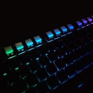 May include: Close-up of a black mechanical keyboard with illuminated keys. The keys are lit with various colors, including green, blue, and purple. The keyboard is angled, showing the function keys and the main keyboard area.