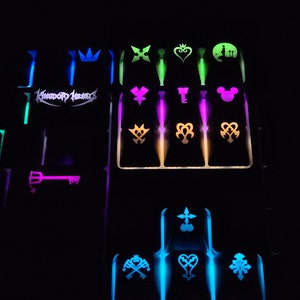 Może przedstawiać: Czarna klawiatura z kolorowymi światełkami i klawiszami z logo Kingdom Hearts i różnymi symbolami z gry. Symbole obejmują koronę, serce, klucz, głowę Myszki Miki i wiele innych unikalnych wzorów.