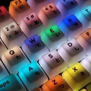 May include: A close-up of a keyboard with colourful backlit keys. The keys have Japanese characters and English letters printed on them. The keys are illuminated in various colours, including red, orange, yellow, green, blue, and purple.