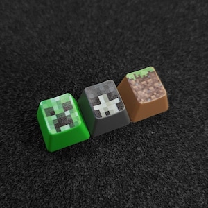 Peut inclure: Trois touches de clavier vertes, grises et brunes avec des motifs sur le thème de Minecraft. La touche verte présente un visage de Creeper, la touche grise présente un bloc de pierre et la touche brune présente un bloc de terre avec de l'herbe sur le dessus.
