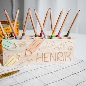 May include: Wooden pencil holder with a light wood finish. The holder has 10 slots for pencils and is decorated with a school theme. The holder is personalized with the name "HENRIK".