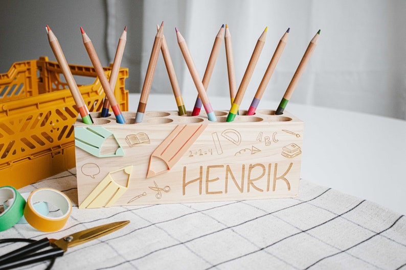 May include: Wooden pencil holder with a light wood finish. The holder has 10 slots for pencils and is decorated with a school theme. The holder is personalized with the name "HENRIK".