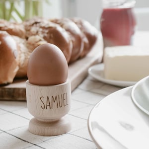 Könnte beinhalten: Ein braunes Ei in einem hölzernen Eierbecher mit dem eingravierten Namen "SAMUEL". Der Eierbecher steht auf einer karierten Tischdecke, mit einem Brotlaib, Butter und einem Glas roter Flüssigkeit im Hintergrund. Eine hellgrüne Schüssel und ein weißer Teller sind im Vordergrund.