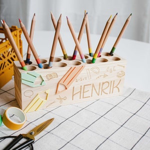May include: A wooden pencil holder with 12 slots for pencils. The holder is decorated with a school-themed design, including a pencil, a ruler, a book, and the letters "ABC". The holder is personalized with the name "HENRIK".