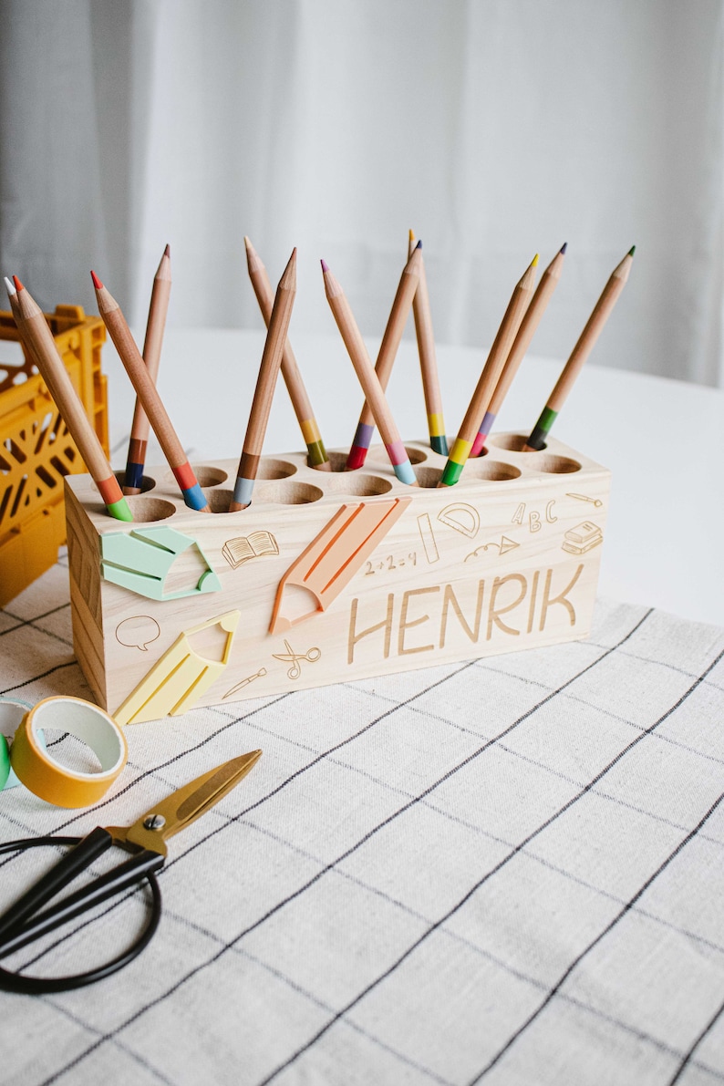 May include: A wooden pencil holder with 12 slots for pencils. The holder is decorated with a school-themed design, including a pencil, a ruler, a book, and the letters "ABC". The holder is personalized with the name "HENRIK".