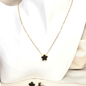 May include: A gold-colored necklace and matching earrings set. The necklace features a delicate chain with a black flower pendant. The earrings are also black flower studs. The jewelry is displayed on a white mannequin.