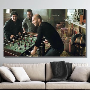 Könnte beinhalten: Ein gerahmter Leinwanddruck zeigt drei Männer, die in einer schwach beleuchteten Bar Tischfußball spielen. Der Tischfußballtisch steht im Mittelpunkt, mit Spielern darum herum. Im Vordergrund sind ein Sofa und eine Pflanze zu sehen.