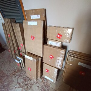 A collection of cardboard boxes and packages wrapped in brown paper, with red shipping labels, stacked against a wall.