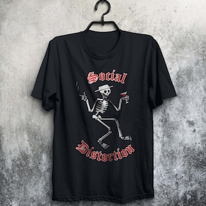 May include: Black t-shirt with a white skeleton wearing a hat and holding a cigarette and a glass of wine. The text "Social Distortion" is printed in red below the skeleton.