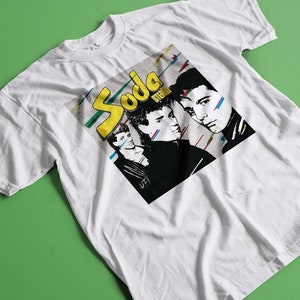 May include: White t-shirt with a graphic print of a band called "Soda Stereo". The band members are depicted in black and white with a colorful background.