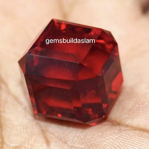 Puede incluir: Una piedra preciosa facetada de color rojo intenso con forma de corte cuadrado y superficie pulida y translúcida. El texto gemsbuildaslam está impreso en blanco en la parte superior central de la piedra, con bordes y facetas geométricas.