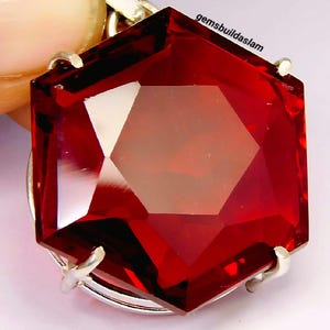 Può includere: Una pietra preziosa esagonale sfaccettata di colore rosso intenso, montata su una montatura in metallo argentato con griffe. La pietra presenta un taglio geometrico prominente con bordi netti e superfici riflettenti. Il testo gemsbuildaslam è stampato in nero vicino al bordo superiore.