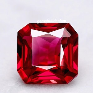 Puede incluir: Una gema de rubí tallada en forma cuadrada, de color rojo intenso. La gema tiene una superficie brillante y reflectante, sobre un fondo blanco. El texto "gemsbuildaslam" es visible en la esquina superior izquierda.