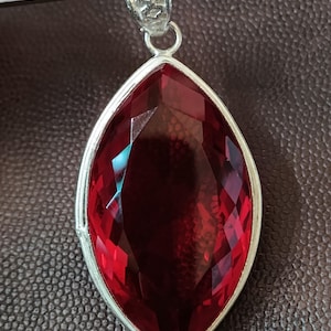 May include: A marquise-cut deep red gemstone pendant set in a silver-toned metal frame. The stone features a faceted surface that reflects light, and the pendant includes a decorative silver-toned bail at the top for attachment to a chain.