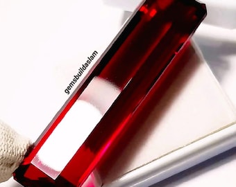 May include: A rectangular, deep red gemstone with a faceted surface and sharp edges. The stone displays high clarity and a vibrant crimson hue. The text 'gemsbuildaslam' is printed in black along the side of the elongated, transparent red crystal.