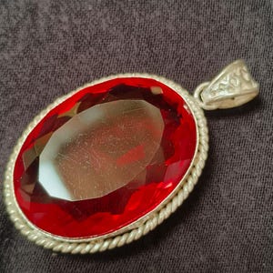 May include: An oval-shaped pendant featuring a large, faceted, deep red gemstone set within a silver-toned metal frame. The frame has a twisted rope-style border and includes a triangular bail at the top for attachment to a chain or cord.