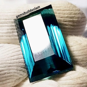 Puede incluir: Una piedra preciosa rectangular facetada con un perfil de color azul verdoso profundo. La piedra presenta una superficie clara y reflectante con bordes afilados y un acabado pulido. El texto "gemsbuildaslam" está impreso en negro.