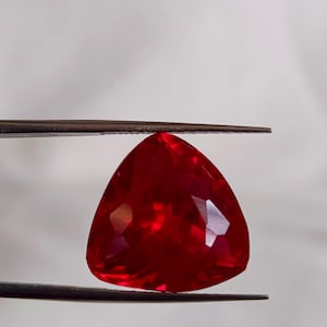 Pode incluir: Uma gema vermelha vibrante, facetada e em forma de triângulo, é segurada por pinças contra um fundo branco. As superfícies polidas da gema refletem a luz, mostrando sua clareza e cor rica. Esta gema é provavelmente uma rubi, adequada para joias.