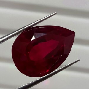 May include: A large, pear-shaped, faceted ruby gemstone. The deep red gem is held by silver tweezers against a white background. The ruby's facets reflect light, showcasing its clarity and brilliance. A close-up view highlights the gemstone's rich colour.