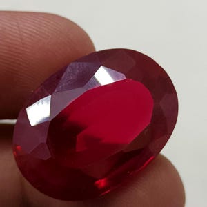 Puede incluir: Una gema de rubí facetada de forma ovalada. La gema es de un color rojo intenso y saturado con múltiples facetas que reflejan la luz. El rubí se sostiene entre los dedos, mostrando su tamaño y brillo.
