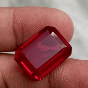 Könnte beinhalten: Ein facettierter, rechteckig geschliffener, tiefroter Edelstein. Der Edelstein hat eine polierte Oberfläche und scharfe Kanten, die das Licht reflektieren. Der Edelstein wird in einer Hand gehalten, um seine Farbe und Klarheit zu zeigen. Der Stein ist wahrscheinlich ein Rubin oder ein ähnlicher Edelstein.