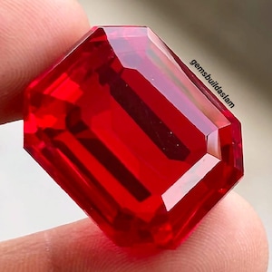 May include: A vibrant, transparent red gemstone with an emerald-cut shape, featuring sharp, clean facets that reflect light. The stone displays a deep, saturated crimson hue throughout its structure. The text gemsbuildslam is visible on the surface.