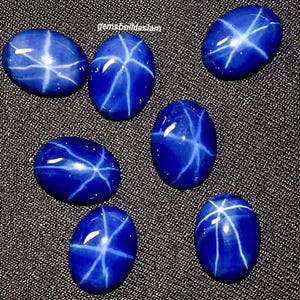 Pode incluir: Sete pedras preciosas ovais, azul profundo, com um padrão de estrela de linhas brancas. As pedras estão dispostas sobre uma superfície escura e texturizada. O texto "gemsbuildasiam" é visível. Adequado para a criação de joias.