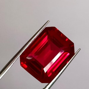 Peut inclure: Un rubis vibrant, de coupe rectangulaire, maintenu par une pince argentée. La pierre précieuse est d'un rouge profond et saturé, avec de multiples facettes réfléchissant la lumière. Le fond est gris neutre.