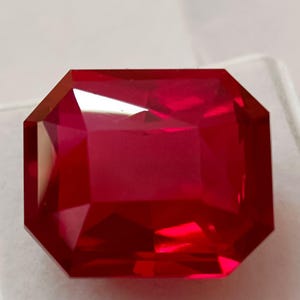 Op de afbeelding: Een gefacetteerde, rechthoekig geslepen, dieprode edelsteen. De edelsteen heeft een briljant, reflecterend oppervlak met meerdere facetten, die de helderheid en kleur laten zien. De edelsteen is geplaatst tegen een neutrale achtergrond.