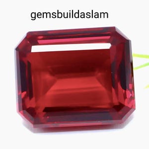 May include: A rectangular, emerald-cut gemstone with a deep, vibrant red colour and faceted surfaces that reflect light. The text 'gemsbuildaslam' is printed in black above the stone, which is positioned against a plain white background.
