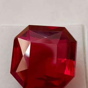 May include: A faceted, rectangular-cut, deep red gemstone. The gem has a complex, multi-faceted surface that reflects light, creating a brilliant display. The gemstone is a rich, saturated red color.